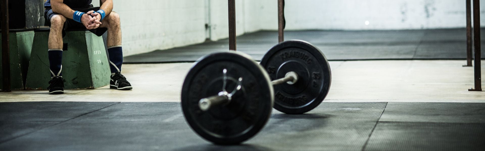 Ao centro, barra de levantamento de peso. No canto esquerdo, pessoa usando roupas apropriadas para levantamento de peso.